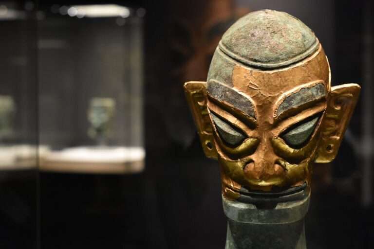 Close-up of an ancient bronze mask exhibited in a dimly lit museum display case.