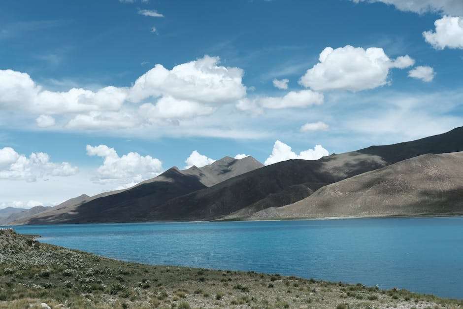 Serene mountain landscape with a crystal-clear lake under a vibrant blue sky and fluffy clouds.