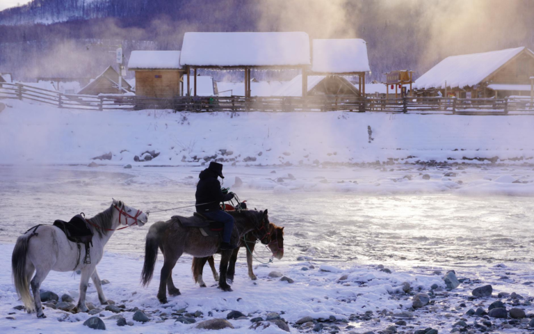 A rider on horseback by a snowy river in Xinjiang during winter with mist rising.