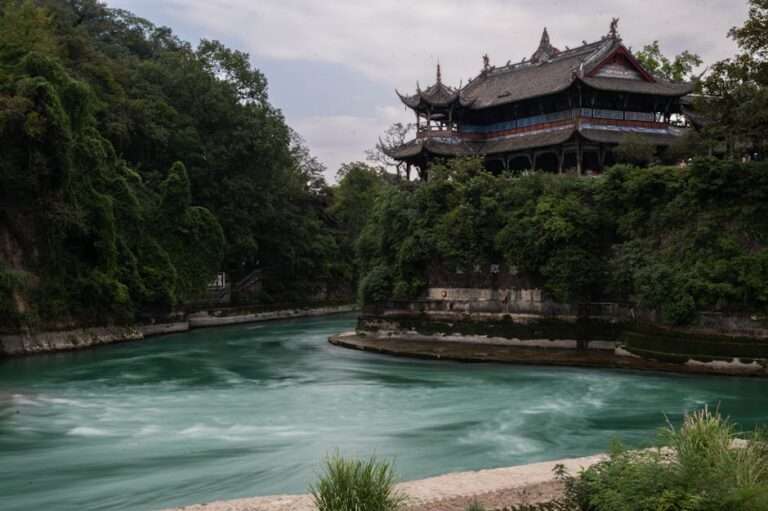 A historic Chinese pavilion surrounded by lush greenery by a vibrant river bend under cloudy skies.