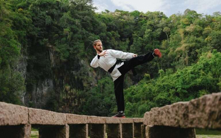 Martial artist in a white uniform balancing on a footbridge in a lush green valley, showcasing fitness and skill.